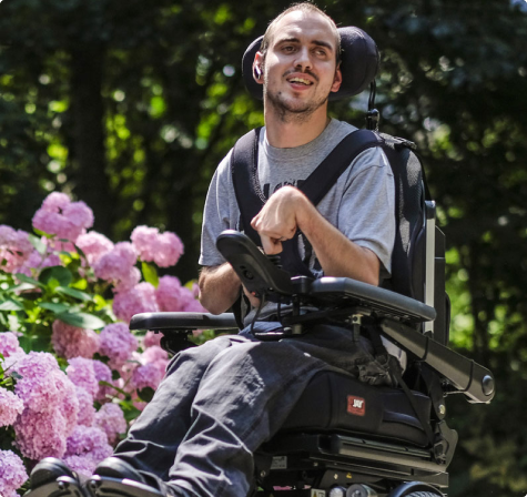 A man sitting in a powered wheelchair outdoors, smiling while surrounded by bright pink hydrangea flowers and leafy trees in the background